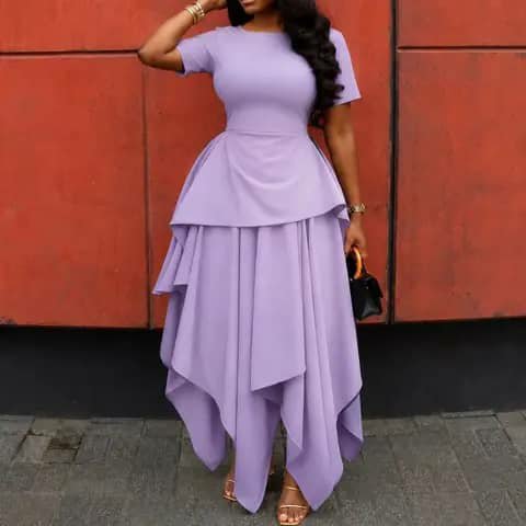 A woman wearing a lilac dress with a peplum top and an asymmetrical skirt, holding a black bag.