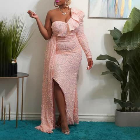 A woman wearing a long pink sequinned one-shoulder dress with a thigh slit, indoors.