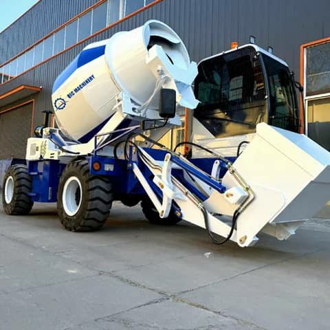 A blue and white self-loading concrete mixer with a front bucket, parked outside.