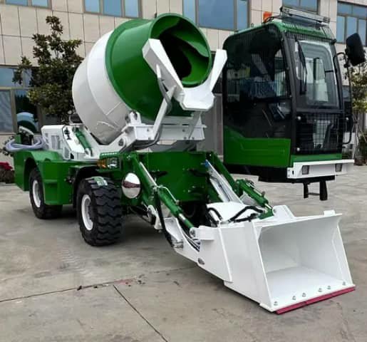 A green and white self-loading concrete mixer with a front bucket, parked outside.